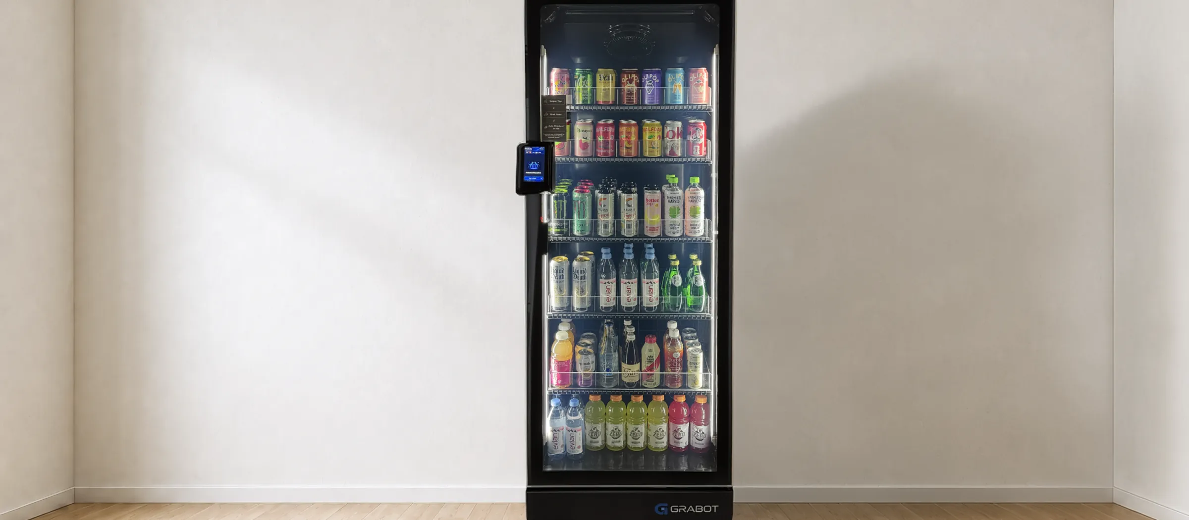 The cooler standing alone against a plain wall in an empty room with a pale wood floor, lit shelves stocked with canned sodas, sparkling waters and bottled drinks.